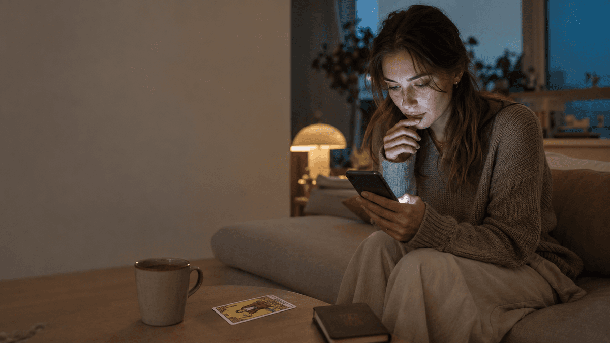 Woman on a sofa at dusk, phone in hand, with one tarot card—The Magician—on the coffee table beside a mug and notebook.